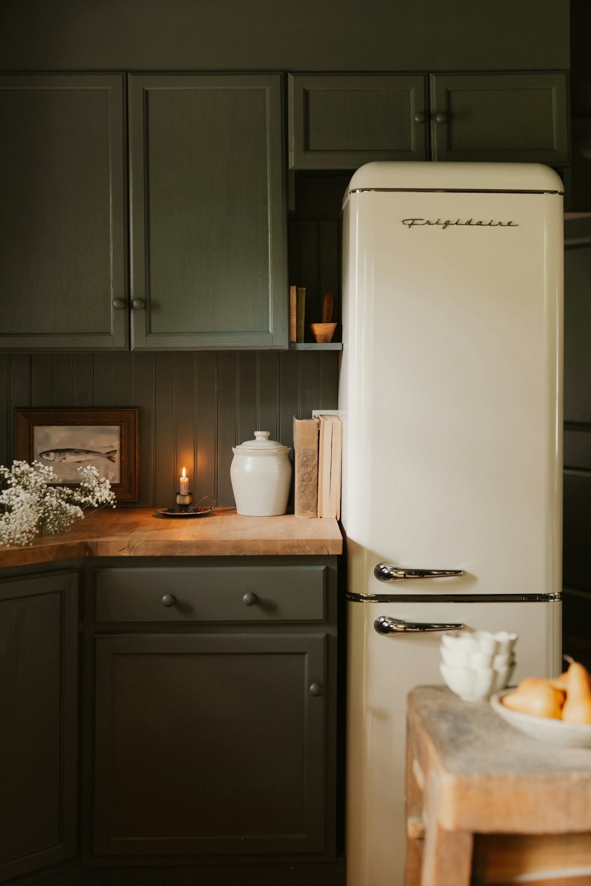 A cream retro refrigerator beside dark green cabinets and a wooden countertop with a candle